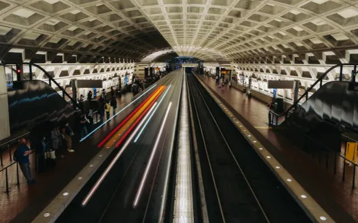 A long-exposure view of a busy underground metro station with a geometric concrete ceiling as a train speeds through the tracks, creating streaks of light while passengers wait on both platforms.